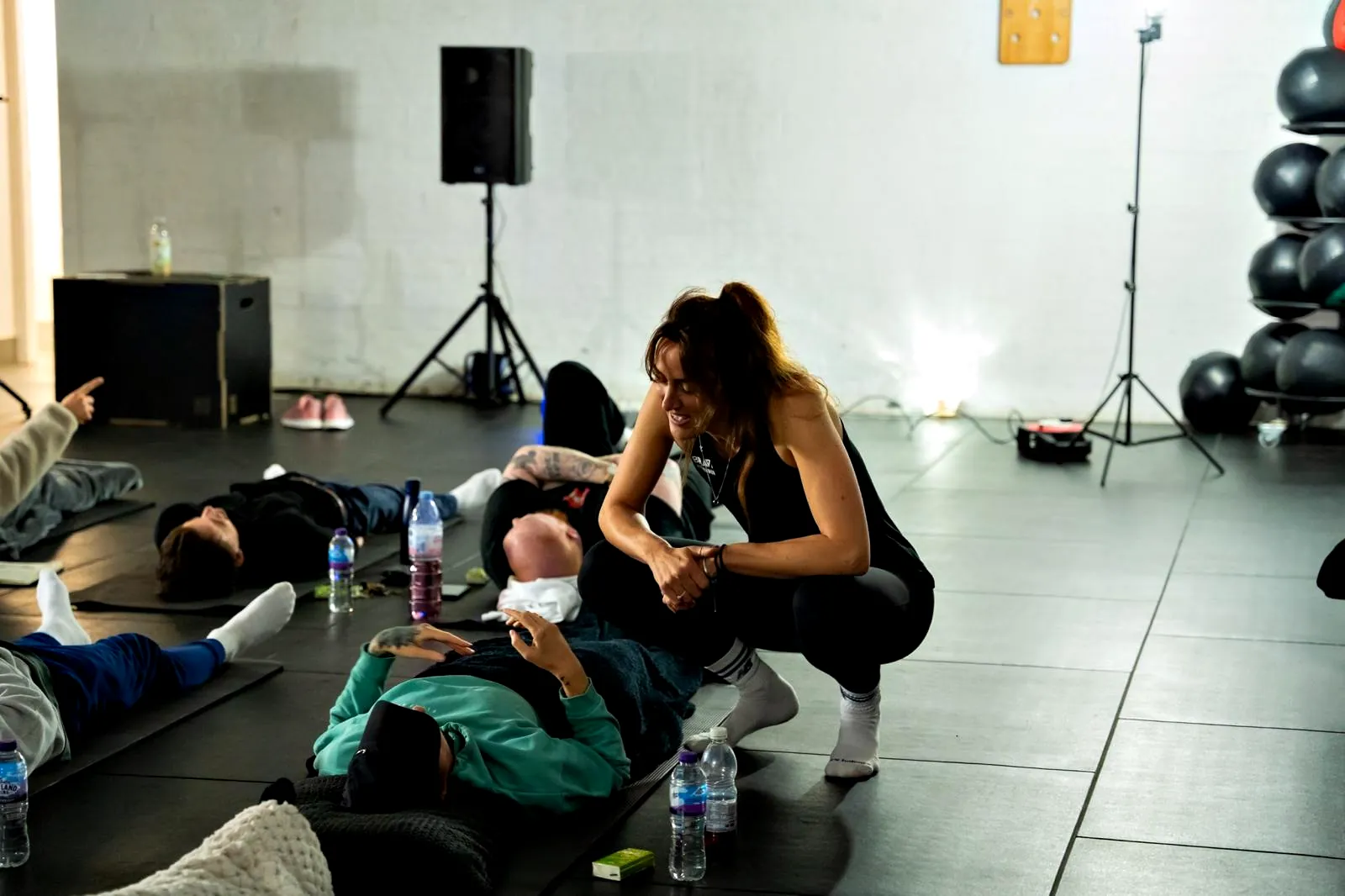 Liane crouching beside someone on their mat during a group session in a gym